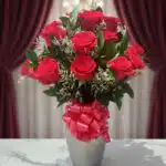 Jarrón plateado con doce rosas rojas frescas y follaje verde, atado con un lazo rojo brillante, sobre una mesa de mármol blan