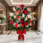 Jarrón de cristal con 18 rosas de varios colores (rojas, blancas, rosas, naranjas) y follaje verde, adornado con un moño rojo