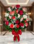 Jarrón de cristal con 18 rosas de varios colores (rojas, blancas, rosas, naranjas) y follaje verde, adornado con un moño rojo