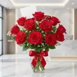 Jarrón de cristal con 18 rosas rojas vibrantes y abiertas, adornado con un elegante moño rojo, sobre una superficie de mármol