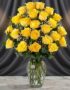 Jarrón de cristal con 24 rosas amarillas frescas y vibrantes, dispuestas elegantemente sobre una mesa de mármol con un fondo