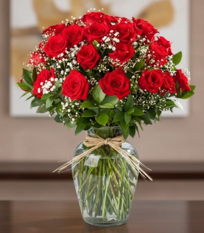 Jarrón de cristal con 24 rosas rojas frescas y follaje verde, atado con un lazo de rafia, sobre una mesa de madera oscura con
