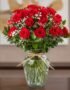 Jarrón de cristal con 24 rosas rojas frescas y follaje verde, atado con un lazo de rafia, sobre una mesa de madera oscura con