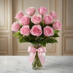 Jarron Allegria con doce rosas rosadas vibrantes y abiertas, adornado con un elegante moño rosa, sobre una superficie de márm