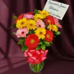 Jarrón de cristal con un vibrante arreglo de gerberas multicolores (rojas, amarillas, rosas) y follaje verde, atado con un la