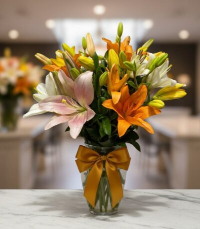 Jarrón de cristal con un moño dorado, lleno de lirios de colores variados: blancos, naranjas, amarillos y rosas, sobre una su