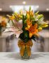 Jarrón de cristal con un moño dorado, lleno de lirios de colores variados: blancos, naranjas, amarillos y rosas, sobre una su