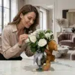 Mujer elegante admira el Jarrón Llévame Contigo con rosas blancas y un osito de peluche, en un interior de hogar de alto dise