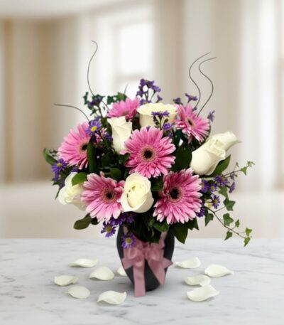 Jarrón Primavera con un elegante arreglo floral de rosas blancas, gerberas rosas y pequeñas flores moradas, sobre una mesa de