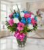 Jarrón Primavera con un arreglo floral vibrante de rosas blancas, gerberas fucsia y alstroemerias azules, sobre una superfici
