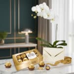 Arreglo MARELY con orquídea Phalaenopsis blanca, caja de chocolates Ferrero Rocher, tarjeta de regalo y cinta dorada sobre má