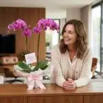 Mujer de mediana edad sonriendo y admirando una Orquídea Premium de Dos Tallos en un elegante interior de hogar con luz natur