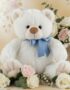 Oso de felpa blanco con lazo azul, rodeado de rosas blancas y rosadas, eucalipto y gypsophila, sobre un fondo elegante y difu