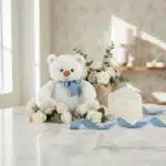 Oso de felpa blanco con lazo azul, rodeado de rosas blancas y rosadas, eucalipto, y una tarjeta de regalo elegante con cinta