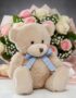 Oso de felpa color miel con lazo azul a cuadros, sentado frente a un elegante ramo de rosas rosadas y blancas, ideal para reg