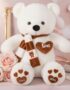 Oso de felpa blanco con bufanda a rayas marrones y blancas, corazón bordado 'LOVE' y patas con huellas de corazón, rodeado de