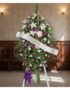 Pedestal fúnebre Esteban con arreglo floral de lirios blancos, rosas moradas y follaje verde, banda blanca con nombre 'Esteba