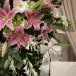 Primer plano de un arreglo floral fúnebre con lirios rosados, margaritas blancas y follaje verde, con rocío sutil y cinta sat