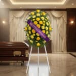 Pedestal fúnebre 'Legado Sereno' con rosas y claveles amarillos, cinta morada 'Sentido Pésame', junto a un ataúd en sala de v