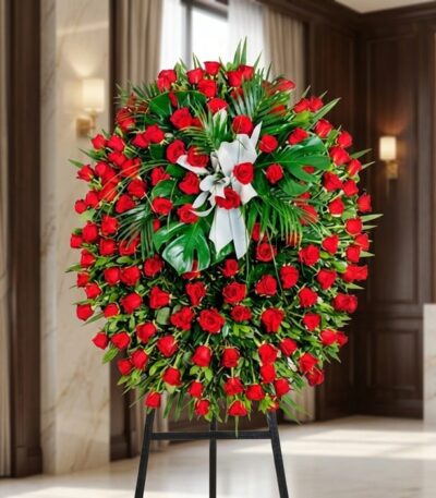 Pedestal fúnebre de rosas rojas intensas y follaje verde, con un lazo blanco, en una sala de velación premium con luz natural