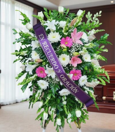 Pedestal fúnebre sagrado con flores blancas, rosas y verdes, con una cinta morada que dice 'En Recuerdo y Oración', junto a u