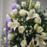 Primer plano de un pedestal fúnebre 'Salvación' con rosas blancas, lirios y follaje verde, destacando texturas y rocío sutil.