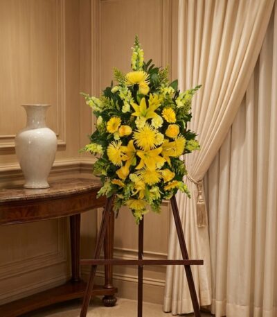 Pedestal de homenaje "El Legado de Sebastian" con un arreglo floral fúnebre de lirios y crisantemos amarillos, presentado ele
