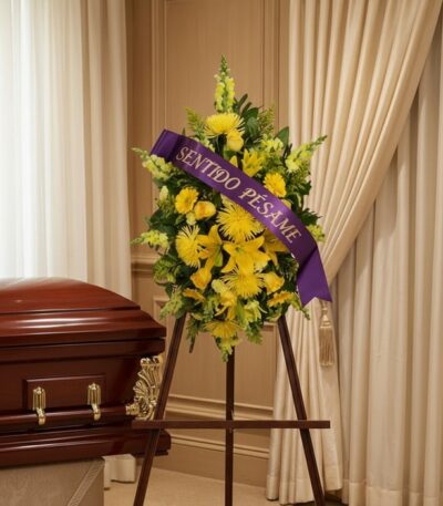 Pedestal de homenaje "El Legado de Sebastian" con flores amarillas y cinta morada "Sentido Pésame" junto a un ataúd en sala d