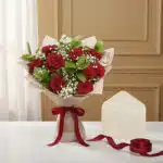 Ramillete de rosas rojas con follaje verde y flores de paniculata, envuelto en papel beige y atado con una cinta de satén roj