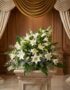 Ramo fúnebre Abdiel con lirios blancos y follaje verde, presentado elegantemente en una sala de velación con cortinas doradas