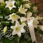 Primer plano de Ramo Fúnebre Abdiel con lirios blancos, follaje verde exuberante y cinta satinada, gotas de rocío sutiles.