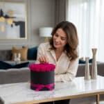 Mujer elegante admirando y tocando delicadamente un arreglo floral de 21 rosas fucsia vibrantes en una caja negra con moño fu
